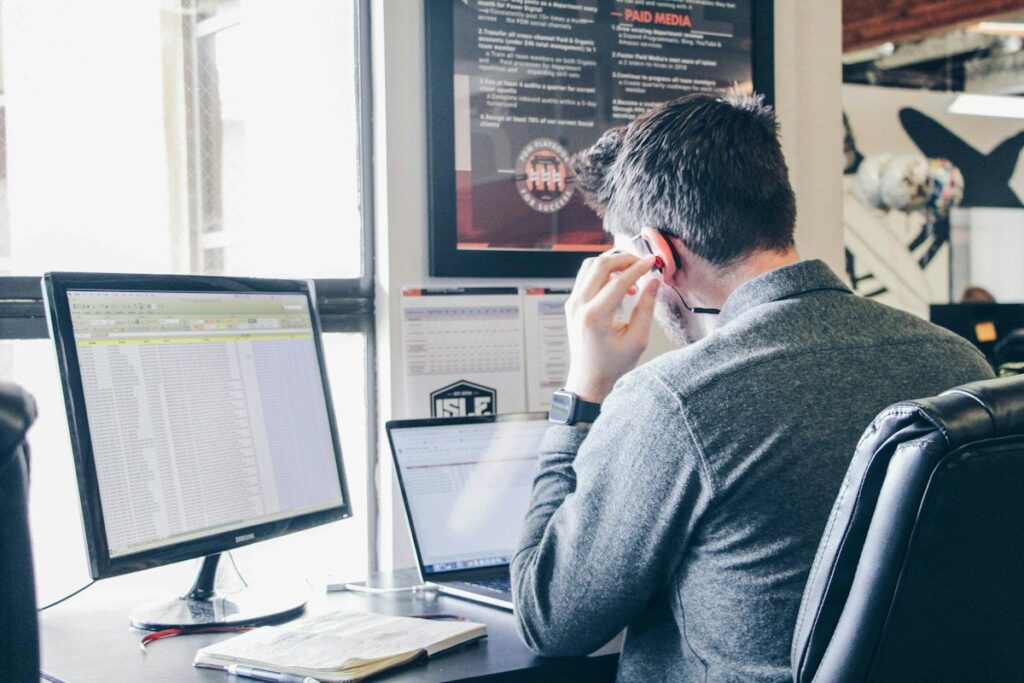 Man at a desk with a spreadsheet on his monitor and a laptop beside him