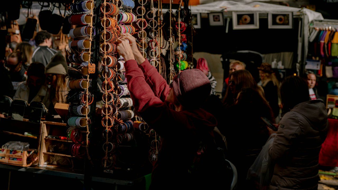Shoppers browsing goods at a busy market stall, where every deal has a buying and a selling price