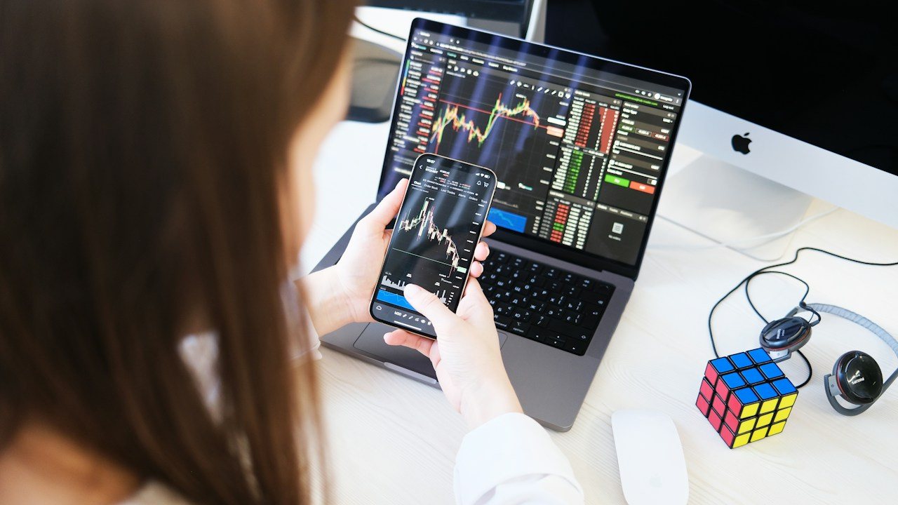 Woman with a phone and a laptop showing trading charts