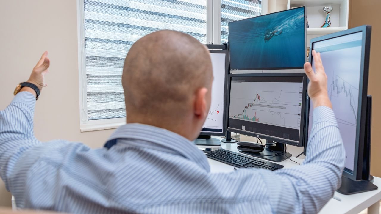 Man with raised arms at a desk with several chart monitors
