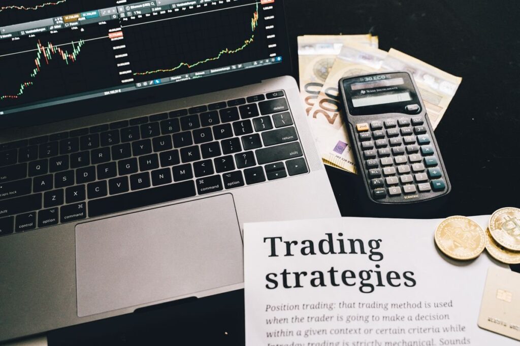 Calculator, coins and a printed trading strategies sheet beside a laptop chart
