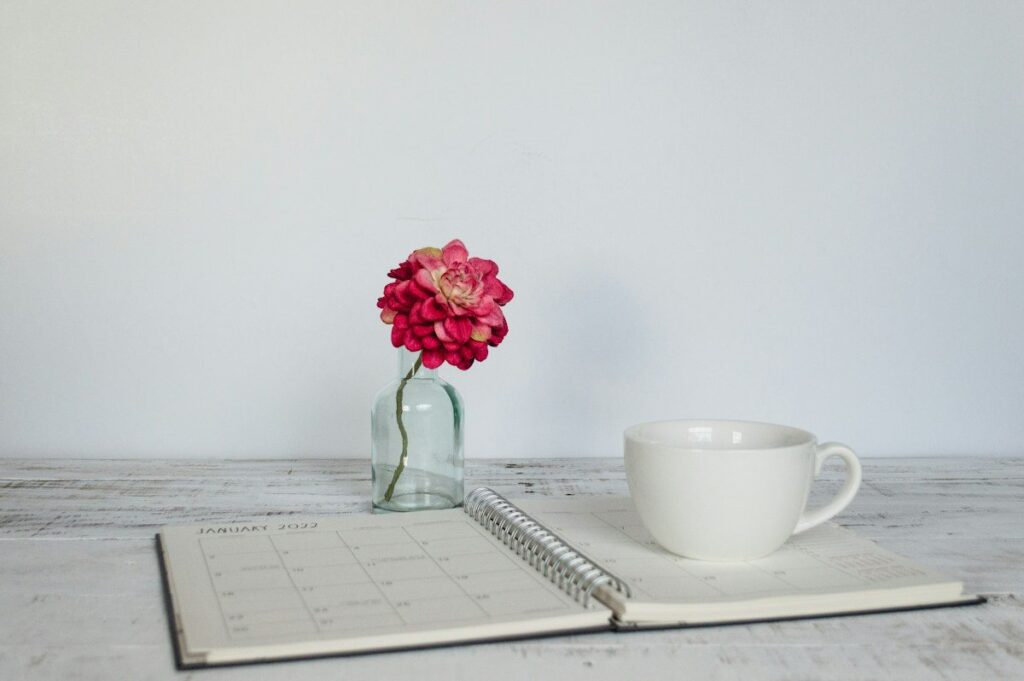 Coffee cup, open planner and a flower in a vase on a white table, part of a fixed morning routine