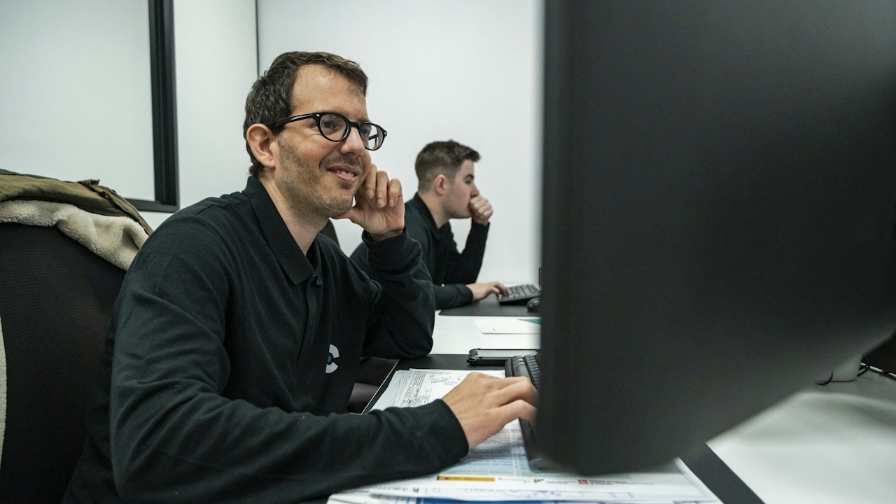 Man smiling at his monitor in an office with a colleague behind him