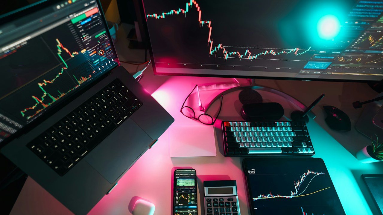 Desk with a laptop and a monitor of candlestick charts, a phone and a calculator under neon light