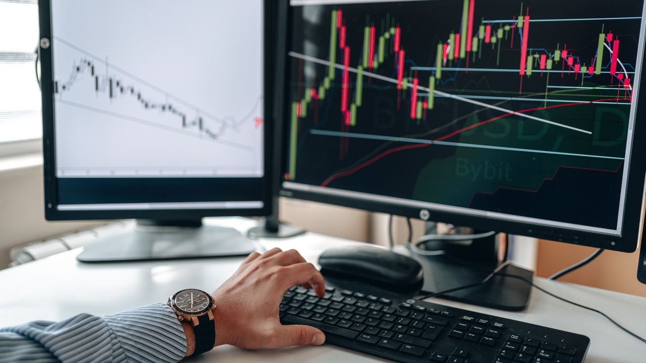 Hand on a keyboard in front of two monitors showing candlestick charts