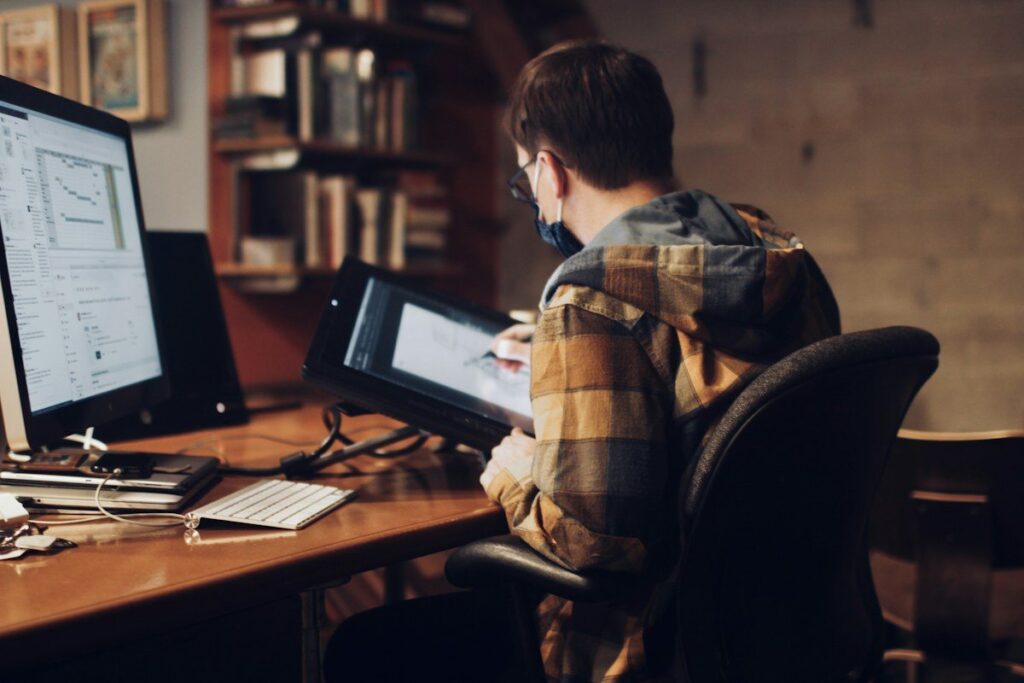 Man in a mask using a tablet at a desk with a monitor
