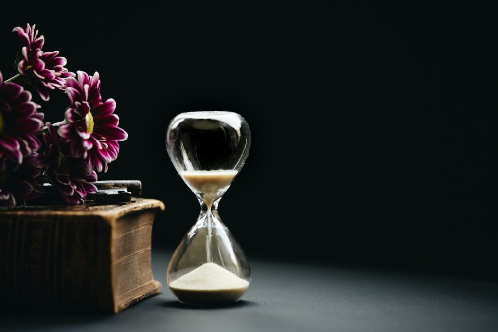 Hourglass beside an old book and flowers on a dark table, representing the long test period a backtest needs to cover