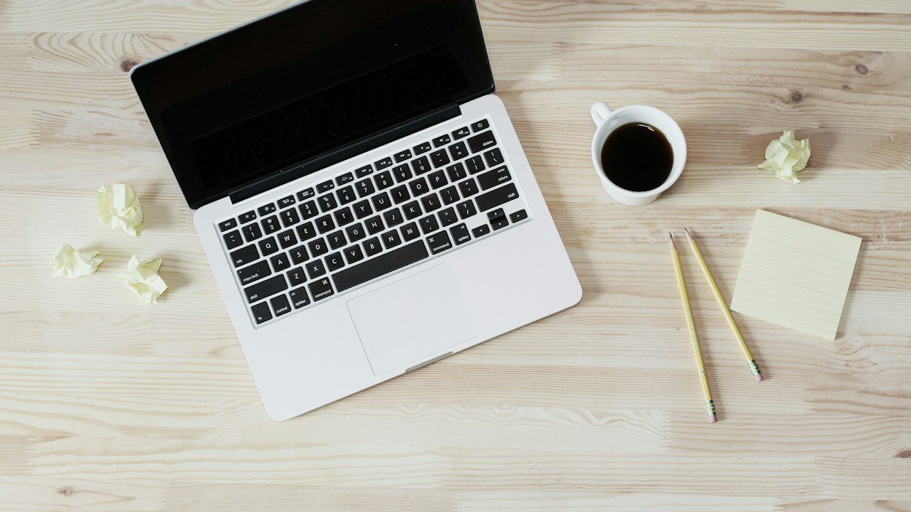 Laptop, coffee and sticky notes on a wooden desk seen from above, the setup where overtrading starts
