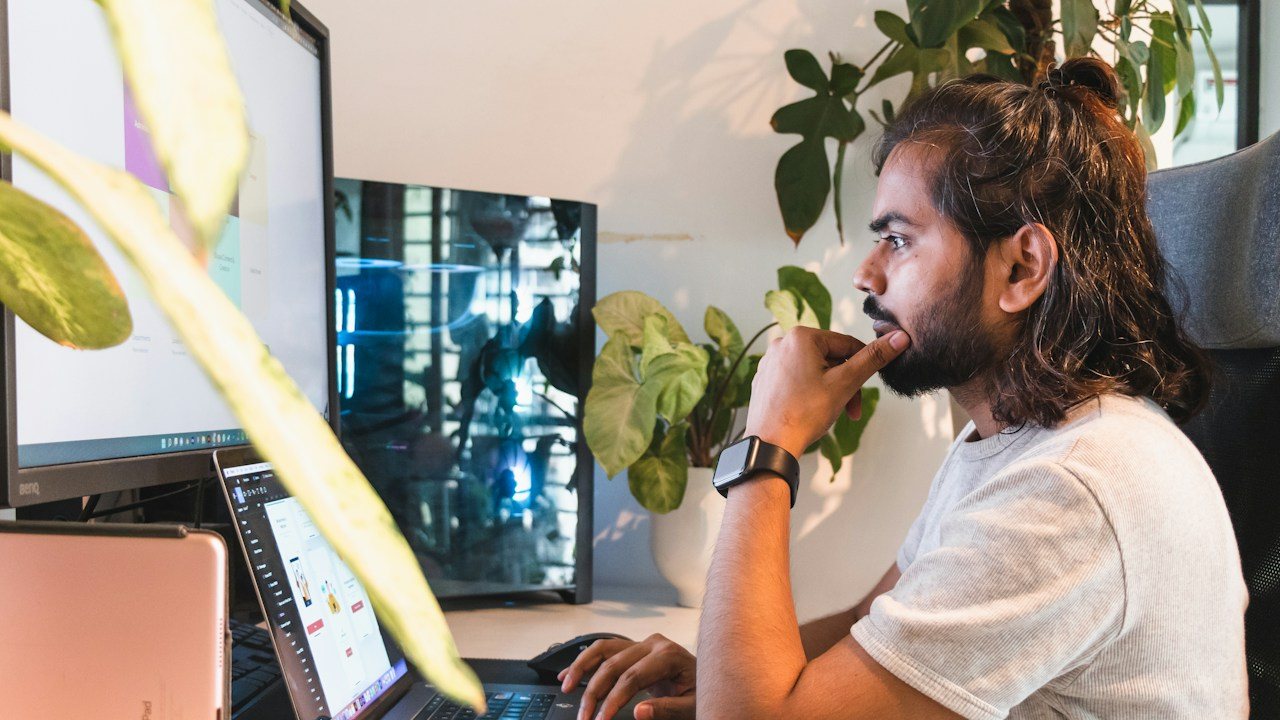 Man at a desk with several monitors, resting his chin on his hand