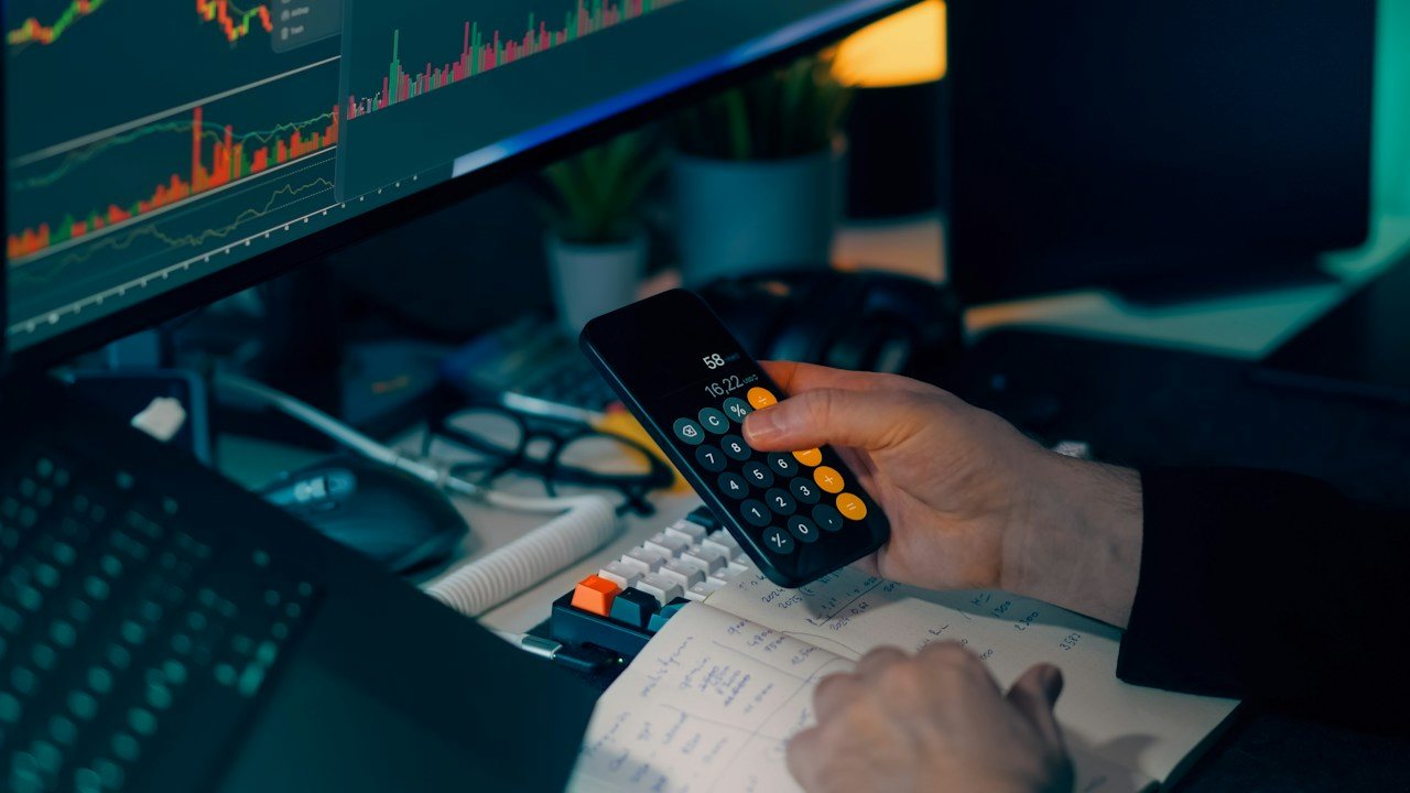 Hand holding a phone calculator in front of a trading chart monitor