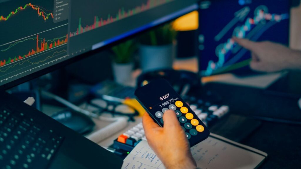 Hand holding a phone calculator in front of trading chart screens