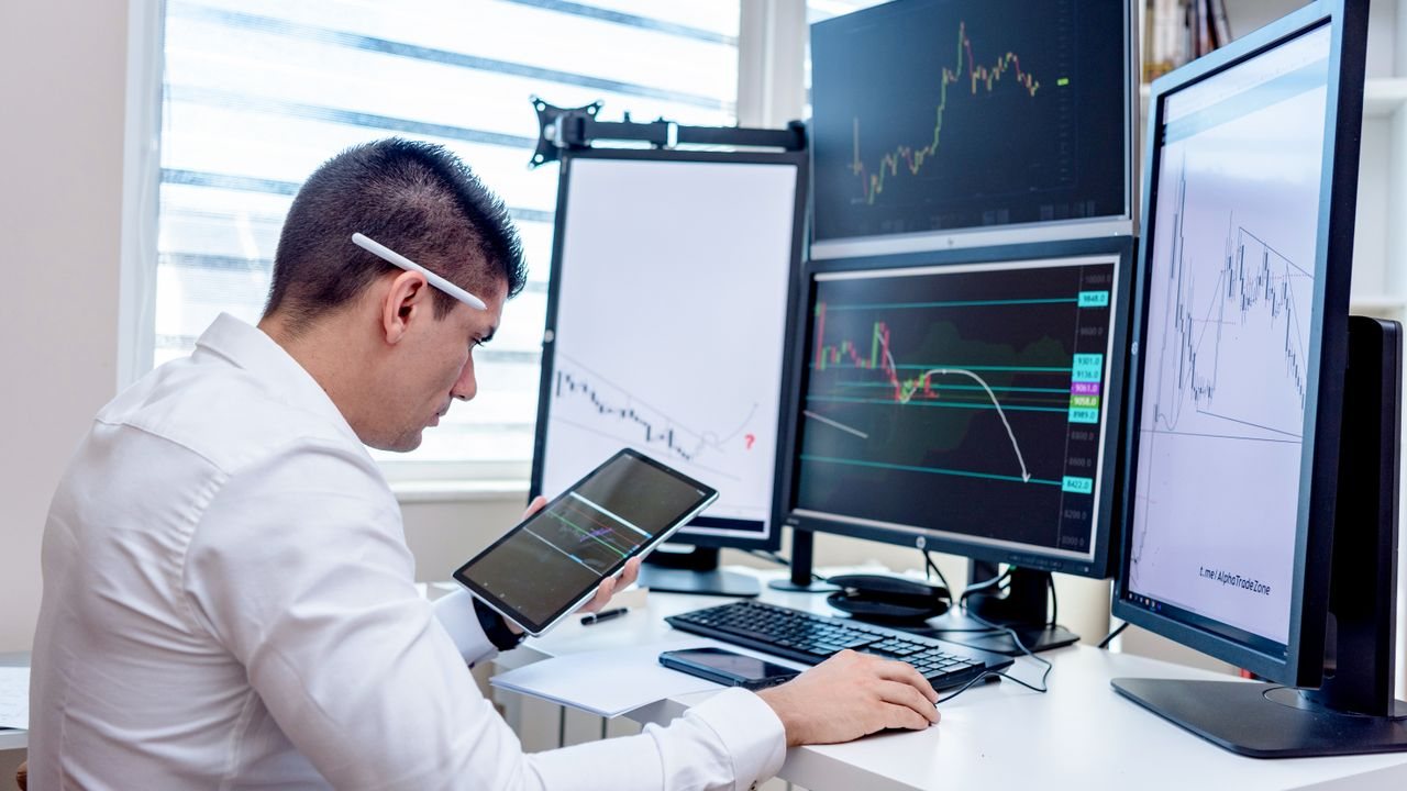Man in a white shirt with a tablet, watching price charts on several screens