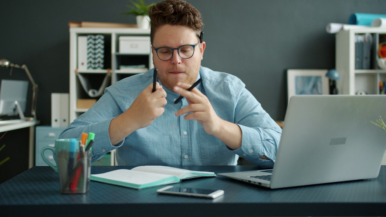 Man at a laptop in a home office, thinking with a pen in his hand