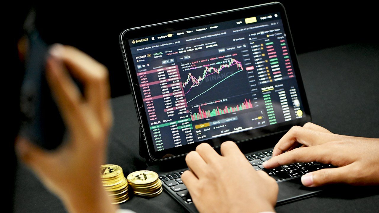 Hands typing on a tablet keyboard showing a trading platform, with coins beside it