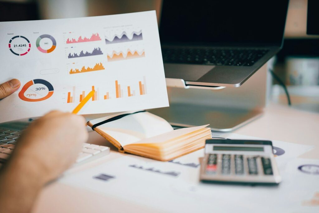 Hand holding a pencil over printed charts beside a calculator and a laptop