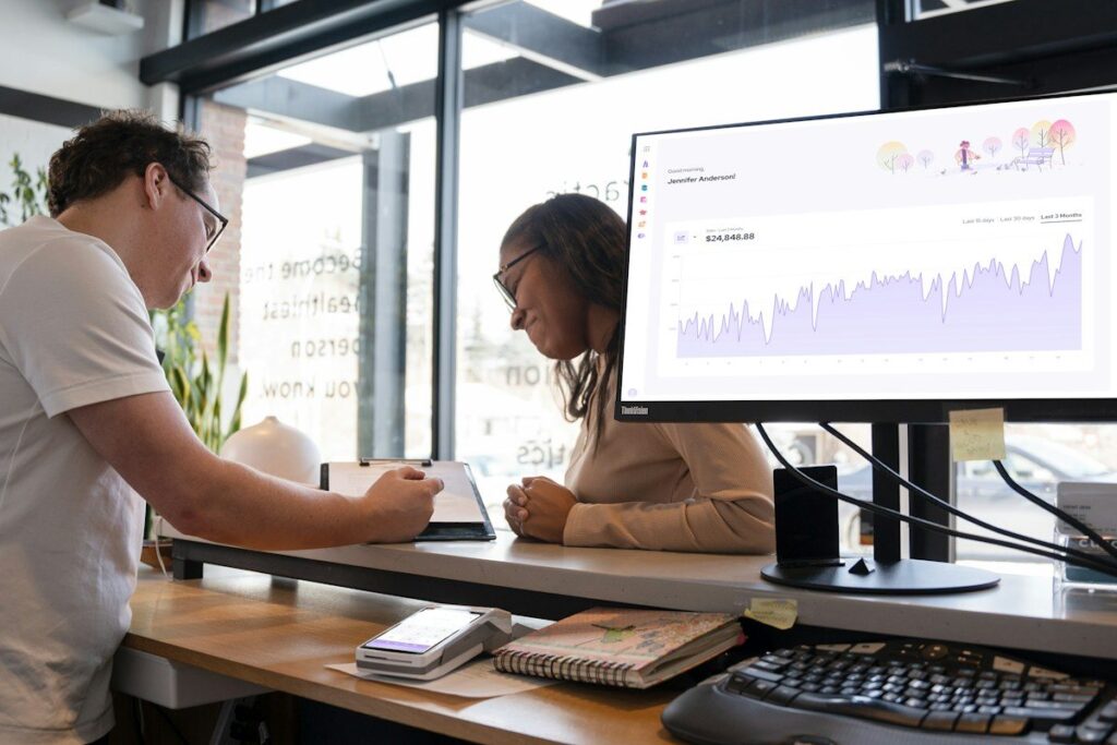 Two people at a desk with a monitor showing a line chart