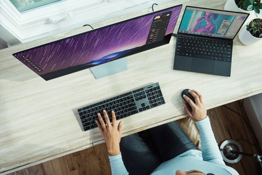 Person at a desk with two computer screens, comparing the FTSE 100, US500, US100 and Germany 40