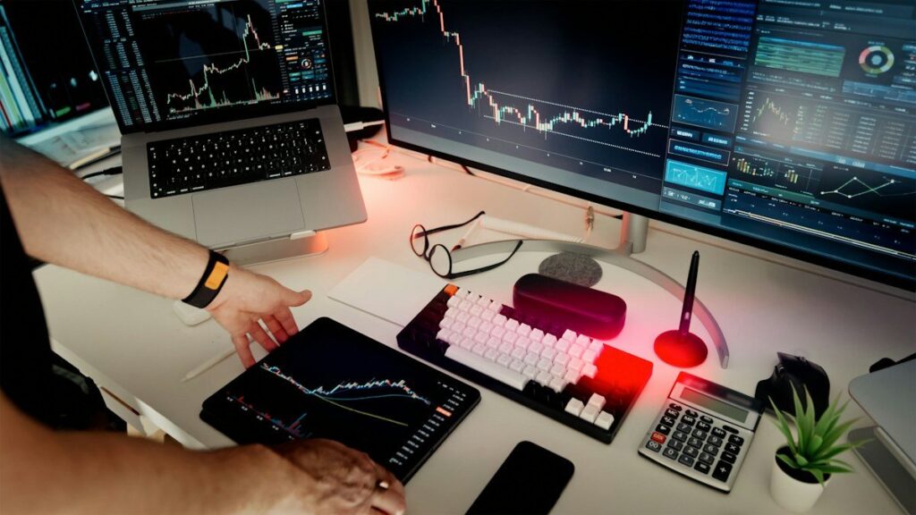 Hands at a desk with several monitors, a tablet chart and a calculator