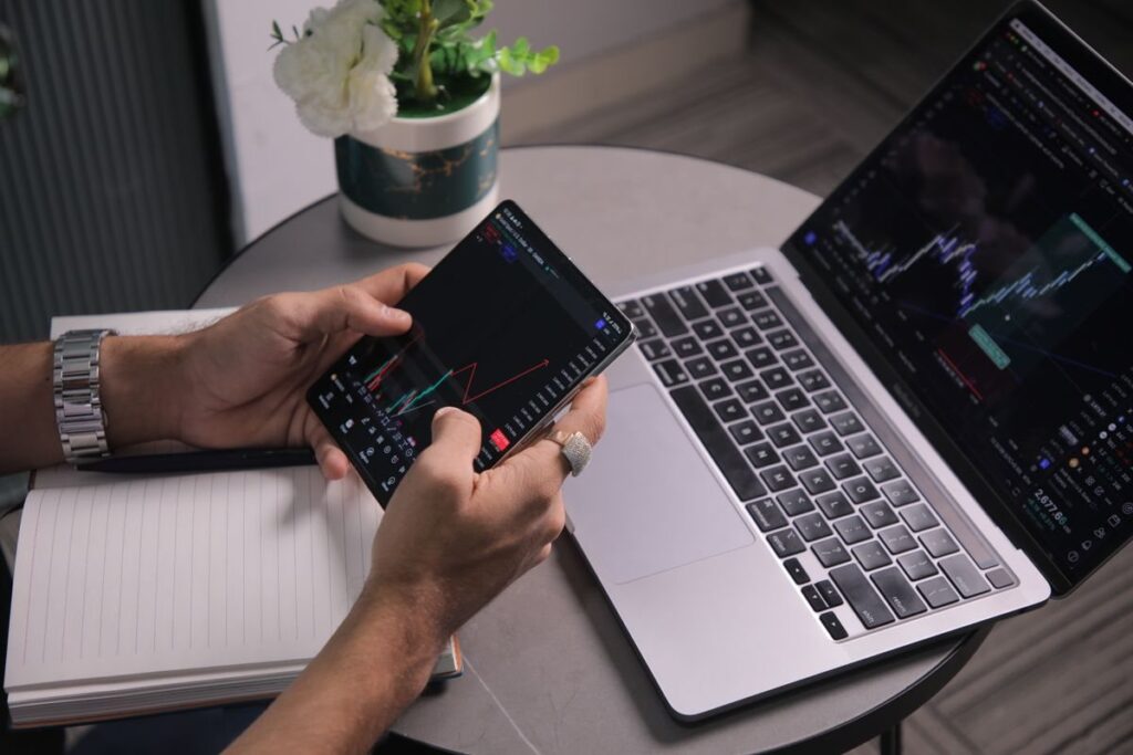 Hands holding a phone with a trading chart beside a laptop and an open notebook