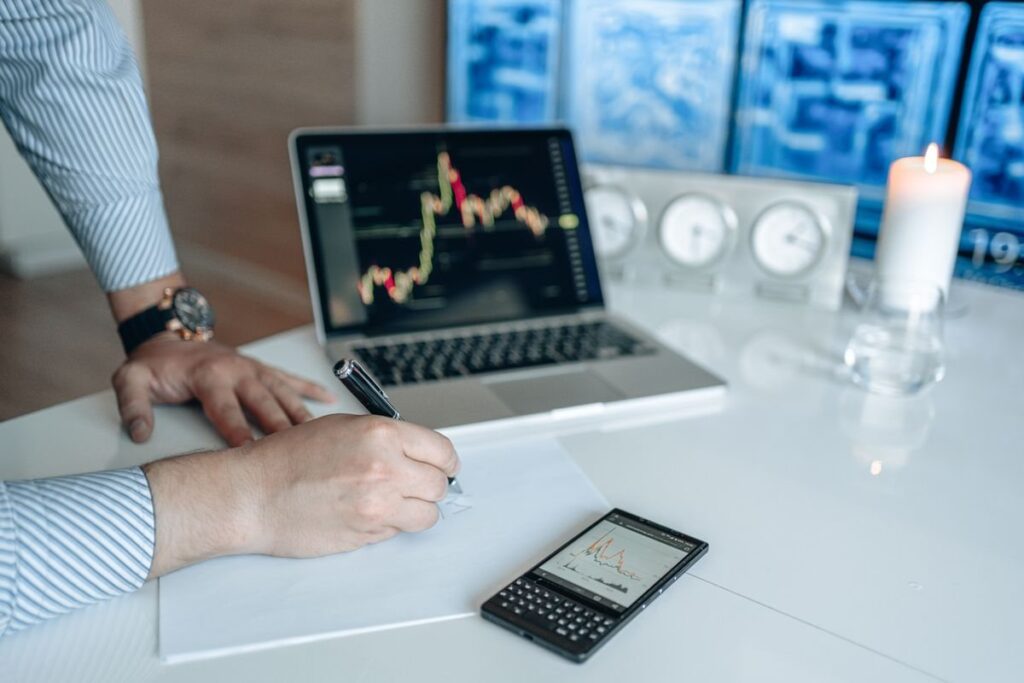 Hand writing beside a laptop showing a candlestick chart