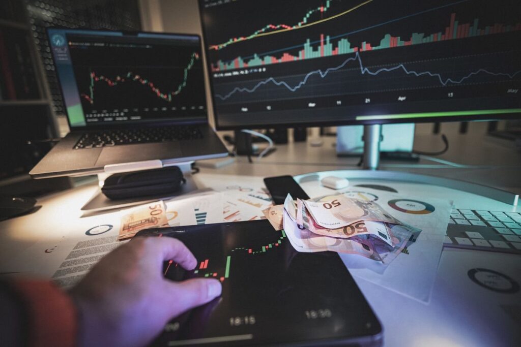 Hand on a phone in front of monitors showing price charts