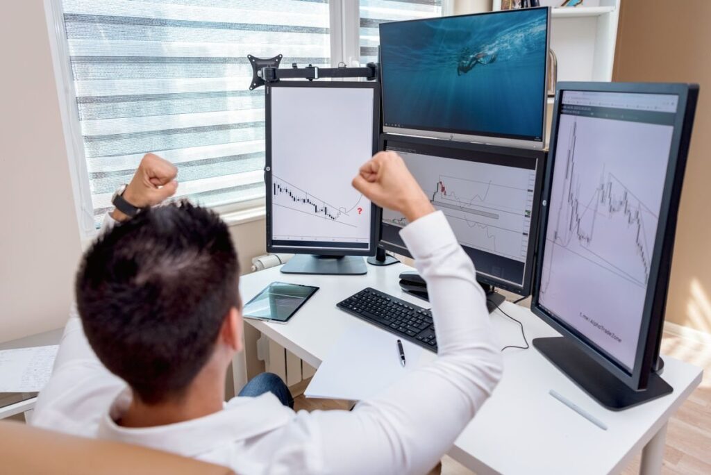 Man cheering at a desk with several chart monitors