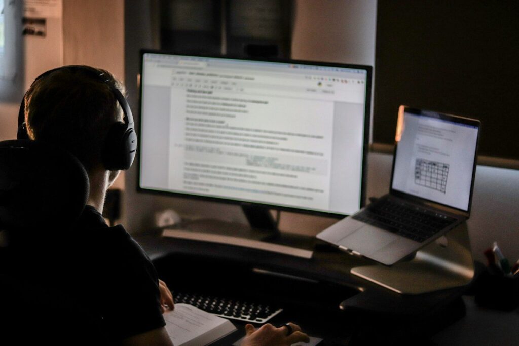 Person at a desk with a monitor and a laptop showing documents and a spreadsheet