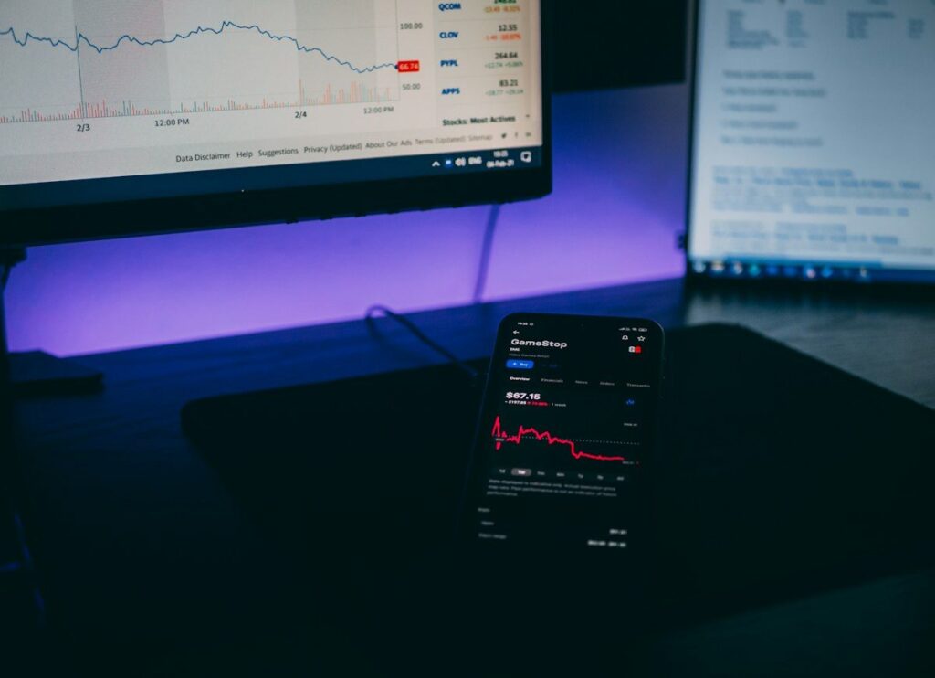 Smartphone with a trading app open beside a computer screen showing a chart
