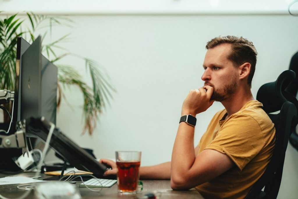 Man in a yellow T-shirt thinking at a desk with monitors