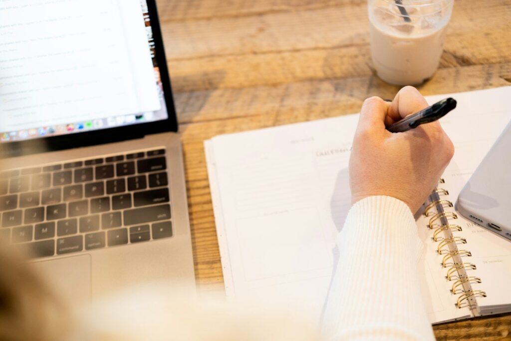 Hand writing in a notebook beside a laptop