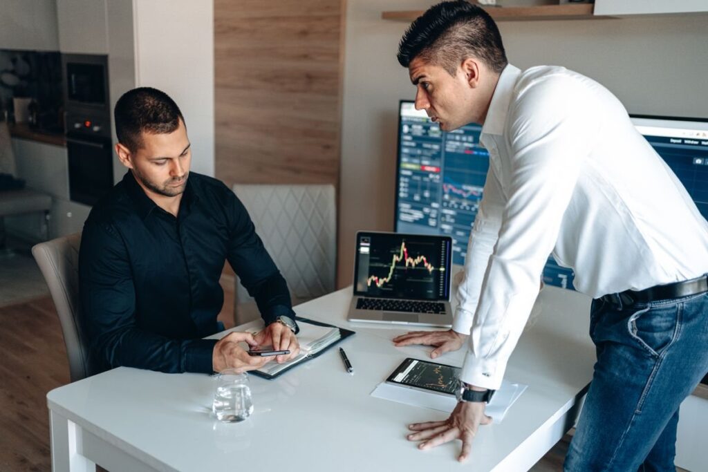 Two men discussing papers beside a laptop chart