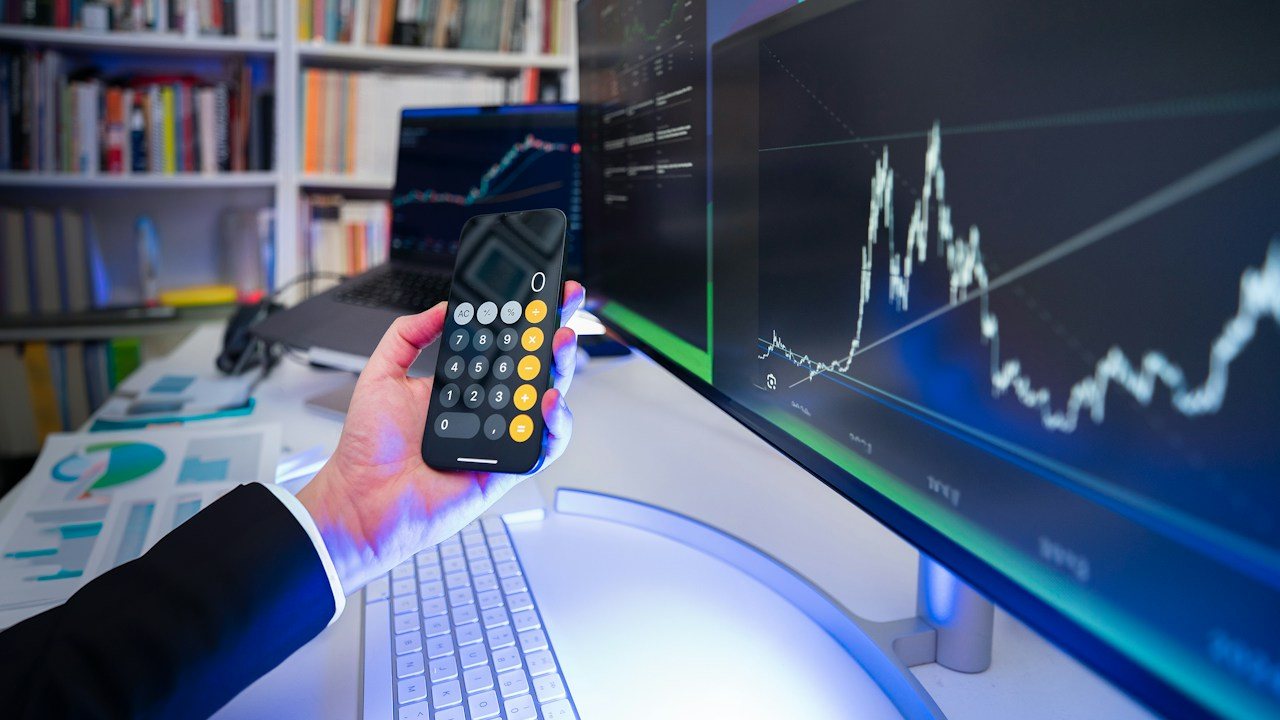Hand holding a phone calculator in front of a monitor showing a price chart