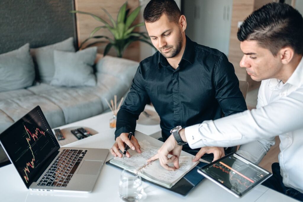 Two men discussing papers beside a laptop chart